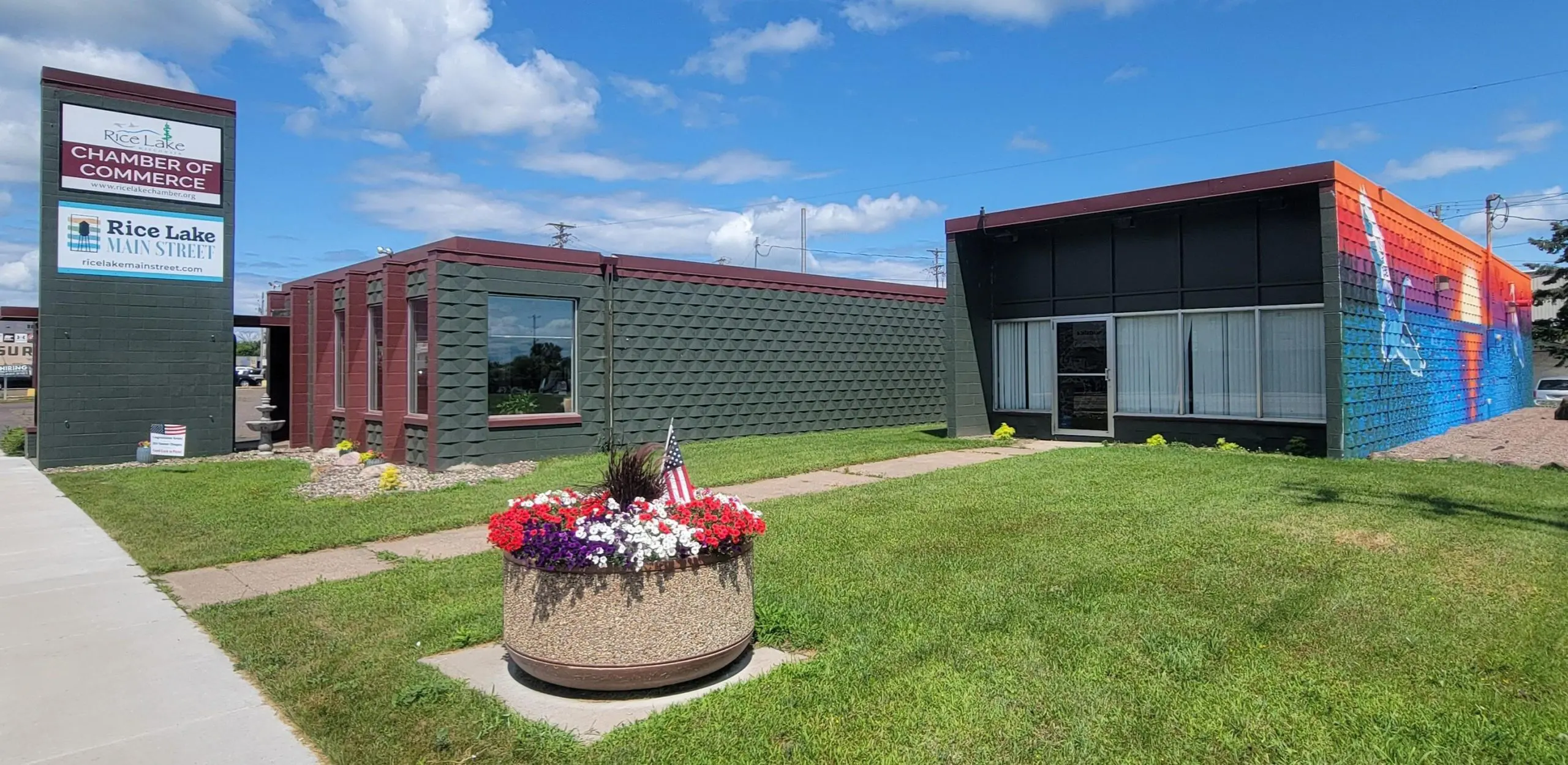 Chamber of Commerce building with a sign for Rice Lake Main Street, featuring a gray and red exterior, and a circular flower planter with American flags in front of a clear blue sky. Our 'About Us' section provides detailed company information and business overview for visitors. - Rice Lake Chamber of Commerce
