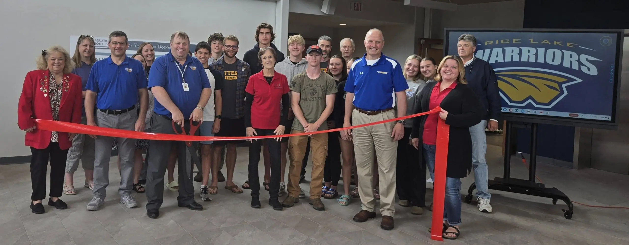 A group gathers in a lobby for a ribbon-cutting ceremony at the Rice Lake Aquatic Center, with a large red ribbon in front and a screen displaying "Rice Lake Warriors" in the background. - Rice Lake Chamber of Commerce