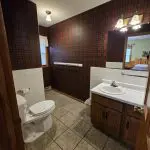 A bathroom at Fred Thomas Resort features plaid wallpaper, a toilet, tiled floor, a white sink with wooden vanity, and a large mirror with lights above it—blending classic style and comfort for over 100 years. - Rice Lake Chamber of Commerce