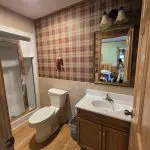 Bathroom with a walk-in shower, toilet, wood vanity with sink, wall-mounted mirror, plaid wallpaper, and hardwood floor—reflecting the timeless style of Fred Thomas Resort as it celebrates 100 years. - Rice Lake Chamber of Commerce