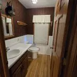 A small bathroom with wood flooring, a white sink and toilet, plaid wallpaper, a window with a blind and valance, and a wooden door partially open—reflecting the cozy charm of Fred Thomas Resort’s 100 years of welcoming guests. - Rice Lake Chamber of Commerce