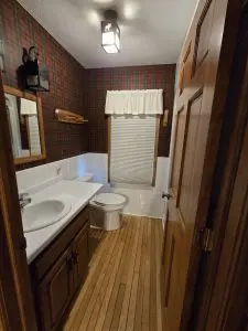 A small bathroom with wood flooring, a white sink and toilet, plaid wallpaper, a window with a blind and valance, and a wooden door partially open—reflecting the cozy charm of Fred Thomas Resort’s 100 years of welcoming guests. - Rice Lake Chamber of Commerce