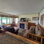 A cozy living room at Fred Thomas Resort with wood-paneled walls, a stone fireplace, rustic furniture, an armchair, a sofa, windows with curtains, and a view of trees outside—celebrating 100 years of comfort and tradition. - Rice Lake Chamber of Commerce