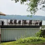 A rooftop deck with lounge chairs and an umbrella overlooks a lake surrounded by trees at Fred Thomas Resort, celebrating over 100 years of welcoming guests. Stairs lead up to the deck from a grassy area below. - Rice Lake Chamber of Commerce
