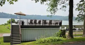 A rooftop deck with lounge chairs and an umbrella overlooks a lake surrounded by trees at Fred Thomas Resort, celebrating over 100 years of welcoming guests. Stairs lead up to the deck from a grassy area below. - Rice Lake Chamber of Commerce