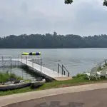 A lakeside scene at Fred Thomas Resort, with a dock extending into the water, several boats on the shore, a gazebo, white chairs, and an inflatable water platform. Trees line the background of this 100-year getaway. - Rice Lake Chamber of Commerce