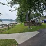 A group of people gather near a small house by a lake at Fred Thomas Resort, with a dock, boats, and trees lining the shoreline, celebrating 100 years of lakeside memories. - Rice Lake Chamber of Commerce