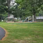 A playground with slides, swings, a gazebo, and nearby basketball hoops sits on a grassy area surrounded by tall trees at Fred Thomas Resort, celebrating 100 years of cherished family memories. - Rice Lake Chamber of Commerce