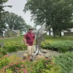 A man in a maroon shirt and khaki shorts stands next to a large fish sculpture at Fred Thomas Resort, surrounded by landscaped gardens, green trees, and buildings in the background—celebrating 100 Years of lakeside tradition. - Rice Lake Chamber of Commerce