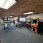 A recreation room at Fred Thomas Resort, featuring a ping pong table, air hockey, two arcade machines, brown couches, and large windows under a wooden ceiling—perfect for enjoying 100 Years of relaxation and fun. - Rice Lake Chamber of Commerce