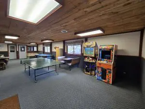 A recreation room at Fred Thomas Resort, featuring a ping pong table, air hockey, two arcade machines, brown couches, and large windows under a wooden ceiling—perfect for enjoying 100 Years of relaxation and fun. - Rice Lake Chamber of Commerce