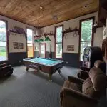 A game room at Fred Thomas Resort with a pool table in the center, three brown recliners, a jukebox, large windows, and shelves with decor celebrates 100 years of relaxation and fun. - Rice Lake Chamber of Commerce