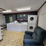 A small laundry room at Fred Thomas Resort features three washing machines, two stacked dryers, a green armchair, wall hooks, a fire extinguisher, and overhead fluorescent lighting—serving guests with comfort for over 100 years. - Rice Lake Chamber of Commerce