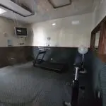 Cozy gym room at Fred Thomas Resort with a treadmill, exercise bike, bench, wall-mounted TV, fan, and carpeted floor. Enjoy beige walls and fluorescent lights—a perfect spot to keep fit during your resort stay. - Rice Lake Chamber of Commerce