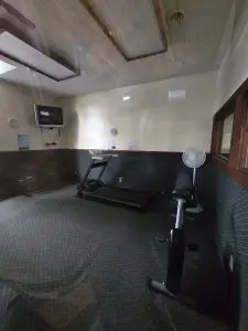 Cozy gym room at Fred Thomas Resort with a treadmill, exercise bike, bench, wall-mounted TV, fan, and carpeted floor. Enjoy beige walls and fluorescent lights—a perfect spot to keep fit during your resort stay. - Rice Lake Chamber of Commerce