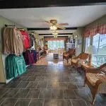 A hallway at Fred Thomas Resort with tile flooring, wicker chairs, a ceiling fan, and a display of colorful sweatshirts and T-shirts on the left wall leads to a windowed seating area—reflecting over 100 years of welcoming guests. - Rice Lake Chamber of Commerce