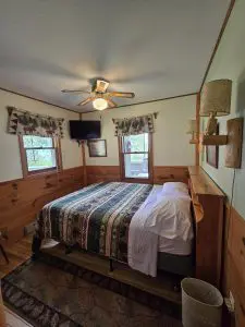 A cozy bedroom at Fred Thomas Resort features a patterned bedspread, wood-paneled walls, a ceiling fan, TV in the corner, and two windows with valances letting in natural light—perfect for celebrating 100 years of relaxation. - Rice Lake Chamber of Commerce