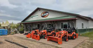 A metal building with Wegman and Olsen's Power & Equipment signs, displaying orange tractors and attachments on a paved area in front of the entrance. Featured in our Member Spotlight. - Rice Lake Chamber of Commerce