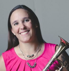 Amanda Kutyaba, a woman with long brown hair, smiles warmly in a pink sleeveless top while holding a brass trumpet. - Rice Lake Chamber of Commerce