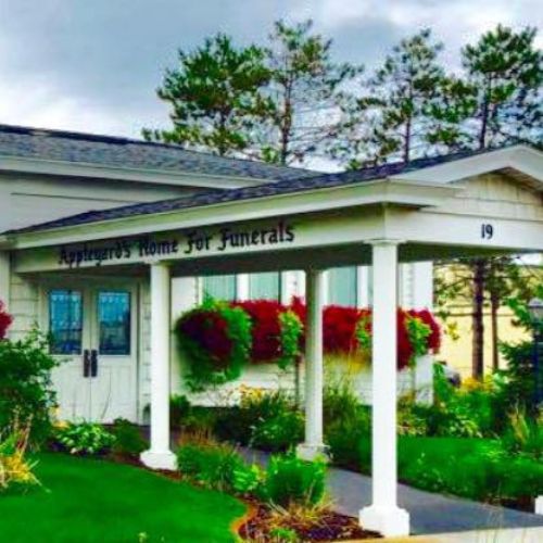 A white building with a sign reading "Appleyards Home for Funerals" stands serenely beside double doors, surrounded by lush greenery and hanging red flowers. - Rice Lake Chamber of Commerce