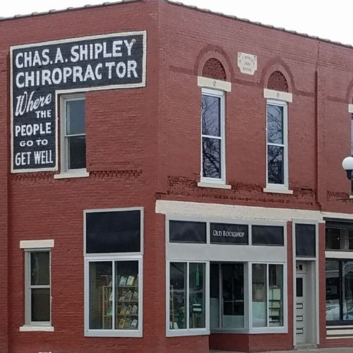 A two-story red brick building in Rice Lake houses a chiropractor's office with a large sign and an old bookshop with a distinctive black and white sign below. The building features elegant white-trimmed windows. - Rice Lake Chamber of Commerce