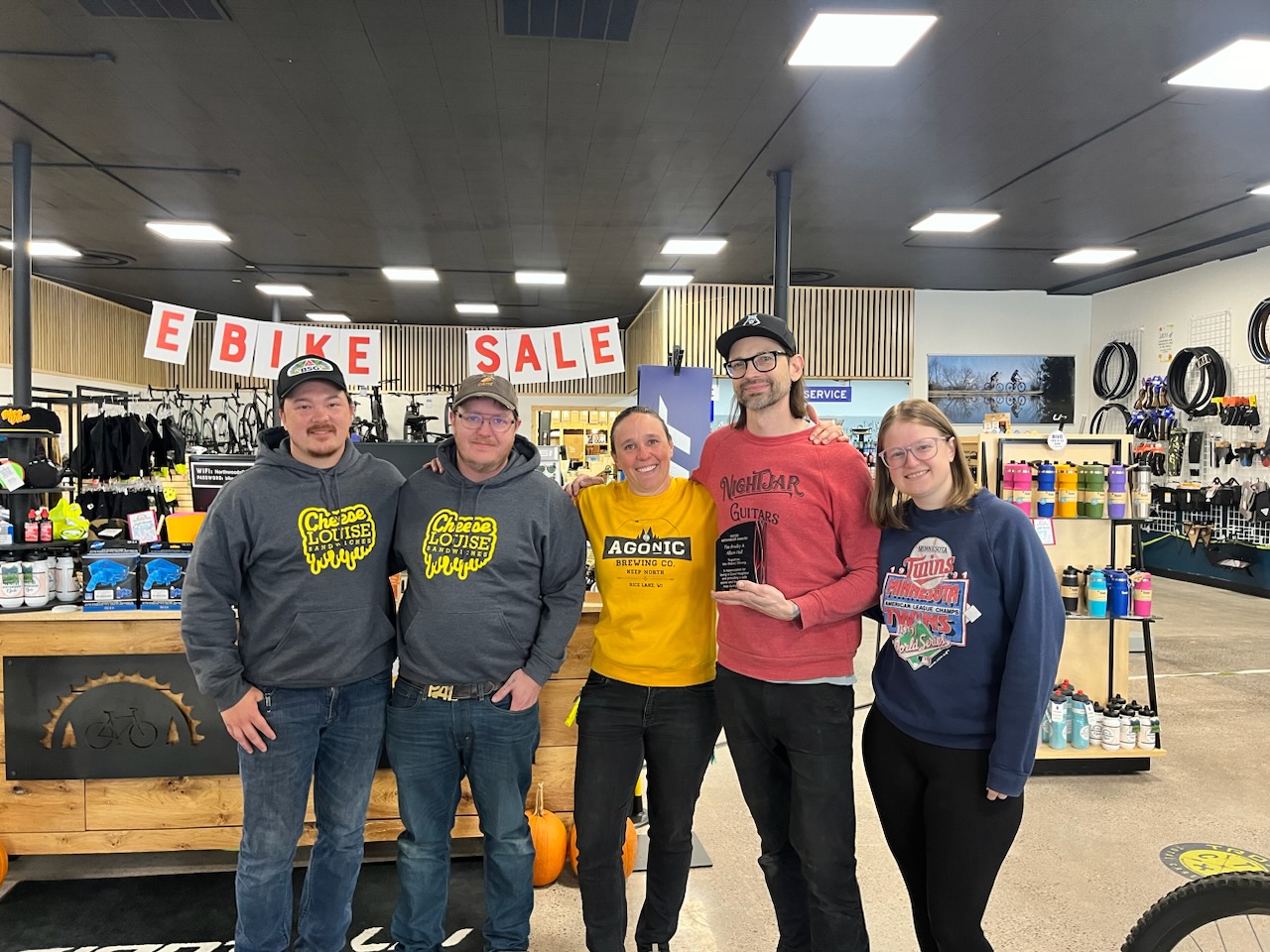 Five people are standing inside Northwoods Cycle, surrounded by bicycles and accessories. They're all smiles, posing for a group photo under an "Ebike Sale" sign, capturing the spirit of our November Member Spotlight. - Rice Lake Chamber of Commerce