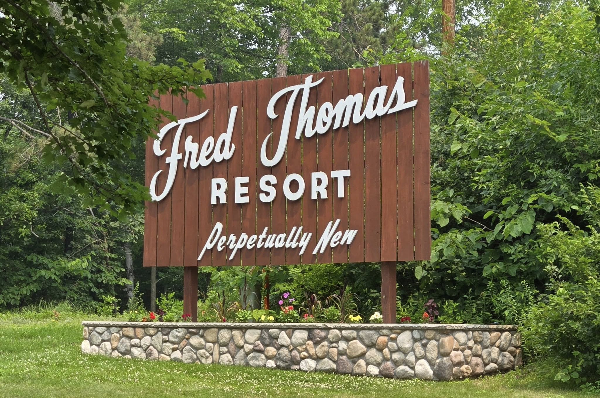 A large wooden sign reads "Fred Thomas Resort Perpetually New," celebrating 100 years, surrounded by trees and a stone-bordered flower bed. - Rice Lake Chamber of Commerce